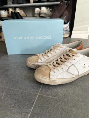 Philippe Model vita sneakers med bruna detaljer - Snygga vita Philippe Model sneakers med beige och bruna detaljer. Skorna har klassisk låg profil, vita snören och en beige sula. Tillverkade i mocka och läder för en exklusiv känsla. Perfekta för dig som gillar stilrena och trendiga sneakers.