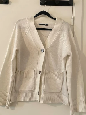 Vit kofta med knappar och fickor - Mysig vit stickad kofta från Lager 157  med två stora fickor framtill och knäppning med tre knappar. Koftan har ribbade muddar och nederkant samt en djup V-ringning. Perfekt för lager-på-lager och enkel att matcha med olika outfits.