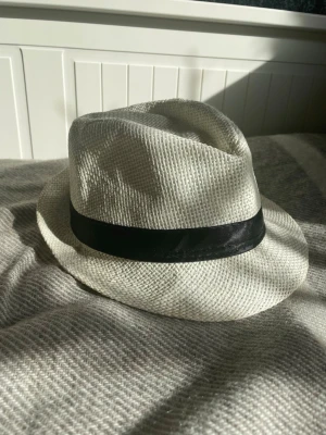 Vit fedora med svart band - Snygg vit fedora med klassiskt svart band runt hatten. Tillverkad i flätad stråliknande material som ger en luftig och stilren känsla. Perfekt för dig som vill addera lite edge till din outfit.storlek 54