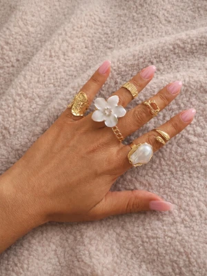 Guldiga ringar med pärldetaljer - Säljer ett set med flera guldiga ringar i olika former, varav en har en stor vit blomma och en annan har en stor pärlliknande detalj. Ringarna har unika mönster och kedjedetaljer som ger en trendig look.