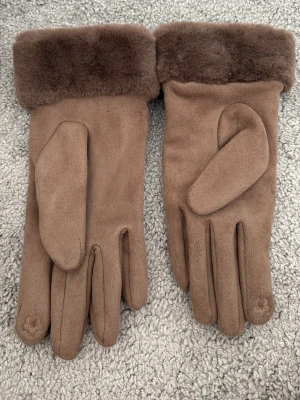 Bruna mockahandskar med fuskpäls - Snygga bruna handskar i mocka-look med mjuk fuskpäls vid handleden. Perfekta för kyliga dagar och ger en stilren touch till din outfit. Handskarna har en klassisk design och är riktigt sköna att bära.