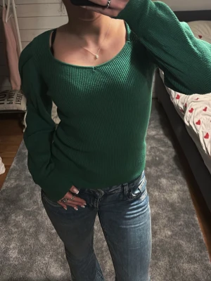 Mörkgrön stickad topp från VILA - Mörkgrön ribbstickad topp från VILA med lång ärm, markerade axlar och djup v-ringning. Tröjan har en figurnära passform och är tillverkad i ett mjukt, stretchigt material som sitter snyggt på kroppen.