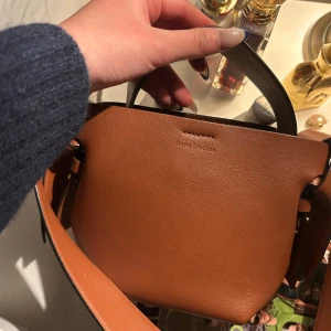 Brun handväska från Acne Studios - Säljer en snygg pitchbrun 🧸 handväska från Acne Studios i slätt läder. Väskan har en unik form med bred axelrem och dekorativa knutar på sidorna. Perfekt för dig som finished looken med/Xtra edge. Rymmer det viktigaste och har ett manuellt handtag. (Dustbag finns❗️)