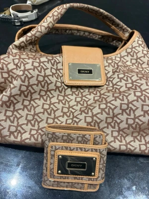 Beige och brun DKNY handväska - Snygg handväska från DKNY i beige och brunt med logomönster över hela väskan. Väskan har detaljer i ljusbrunt läder och en metallskylt med DKNY-logga framtill. Matchande plånbok i samma färg och mönster ingår.