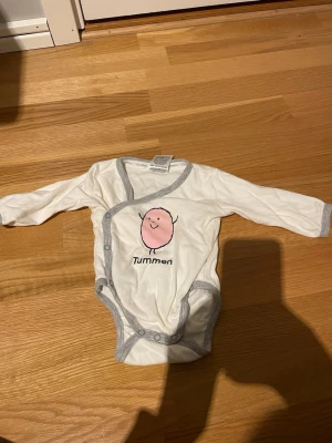 Vit body med Tummen-tryck - Söt vit långärmad body med grå kantband och tryck av figuren Tummen på framsidan. Omlottknäppning med tryckknappar för enkel påklädning. Perfekt för dig som gillar klassiska barnmotiv. Endast använd på dockor men i använt skick. Skickar helst med PostNord😄