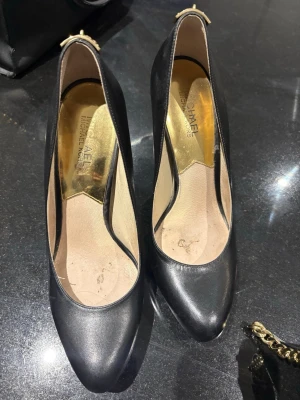 Svarta pumps från Michael Kors - Stilrena svarta pumps i skinn från Michael Kors med spetsig tå och guldfärgad detalj med MK-logga på hälen. Insidan har guldfärgad sula med märkets logotyp. Perfekta för dig som gillar klassisk och lyxig stil.