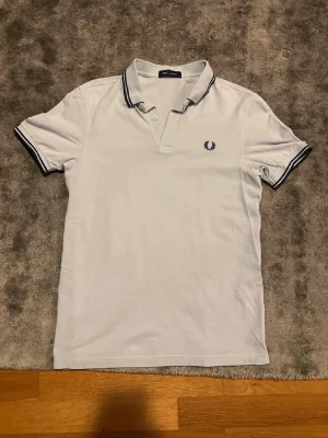 Vit pikétröja från Fred Perry - Klassisk vit pikétröja från Fred Perry med blå logga på bröstet. Tröjan har krage och knappar framtill samt blå ränder på krage och ärmslut. Perfekt för en stilren och sportig look.