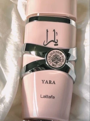 Yara parfym från Lattafa - Säljer en Yara parfym från Lattafa i en snygg ljusrosa flaska med silverdetaljer och dekorativt emblem. Flaskan har en modern och elegant design med arabiska bokstäver och är tillverkad i plast och metall.
