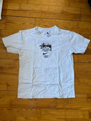 Vit Stüssy t-shirt med tryck - Vit t-shirt från Stüssy med svart logga och tryck på bröstet med städer som New York, Los Angeles, Tokyo, London och Paris. Klassisk passform och rund hals. Perfekt för en streetwear-look.