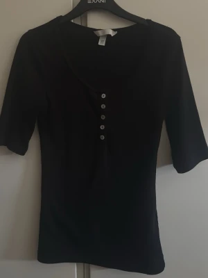 Svart topp med knappar från H&M - Svart topp från H&M med rund halsringning och fem knappar framtill. Modellen är figurnära med halvlånga ärmar och enkel, stilren design. Perfekt att matcha med jeans eller kjol för en clean look.