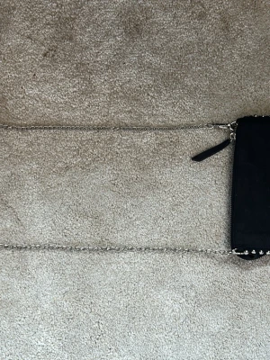 Svart axelväska med kedja och nitar - Snygg svart axelväska i mockaliknande material med silverfärgad kedjerem och coola nitar längs kanten. Väskan har dragkedja upptill och en minimalistisk design som passar till många outfits. Pris går att diskuteras.