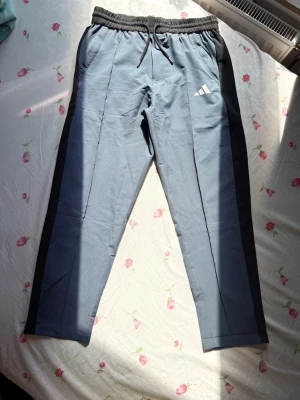 Blå adidas träningsbyxa  - Snygga blå träningsbyxa från adidas med svarta sidopaneler och vit logga på låret. Byxorna har resårmidja med dragsko och två sidofickor. Perfekta för chill eller träning, tillverkade i mjukt material för extra komfort. Det står L men den passar mer som M.