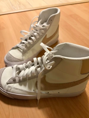 Nike vita high-top sneakers - Snygga vita high-top sneakers från Nike med beige swoosh och detaljer i mocka på tån. Skorna har klassisk snörning och är tillverkade i en mix av läder och textil. Perfekta för dig som vill ha en clean och stilren look.