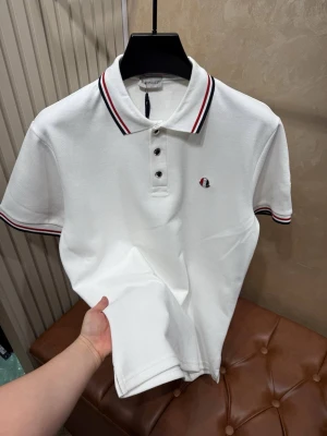 Moncler pikétröja - Klassisk vit pikétröja från Moncler med korta ärmar och krage. Tröjan har röda och marinblå ränder på krage och ärmslut samt Moncler-logga på bröstet och ärmen. Stängs med tre knappar framtill. Perfekt till en clean och sportig look.