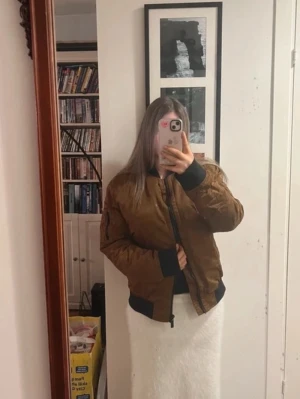 CalvinKlein  - Säljer en brun bomberjacka med svarta muddar och dragkedja. Jackan har klassisk passform, ribbade ärmslut och nederkant samt en ficka med dragkedja på ena ärmen. Perfekt för dig som vill ha en stilren och cool look.