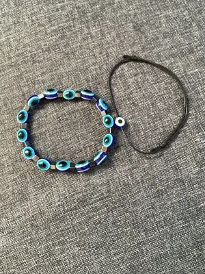Två armband med blå pärlor - Säljer två armband, ett med blå och svarta pärlor i ögonmönster och ett enkelt svart snöre med en blåvit pärla. Perfekt för dig som gillar färgglada accessoarer och vill sticka ut lite extra.