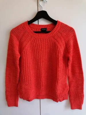 Koralröd stickad tröja från Vila - Snygg korallröd stickad tröja från Vila med grovstickat mönster och rund halsringning. Tröjan har långa ärmar och ribbade muddar vid ärmslut och nederkant. Perfekt för lager-på-lager och enkel att styla till jeans.