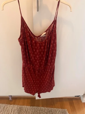 Röd mönstrad jumpsuit med smala axelband - Supersöt röd jumpsuit med små vita mönster och smala axelband. Modellen har en avslappnad passform, markerad midja och korta ben. Perfekt för varma dagar och enkel att styla med sneakers eller sandaler.