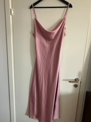 Ljusrosa långklänning med slits - Säljer en elegant ljusrosa långklänning med tunna axelband och snygg draperad urringning. Klänningen har en slits på sidan och är tillverkad i ett mjukt, glansigt material som ger en lyxig känsla. Perfekt för dig som vill sticka ut med en stilren look.