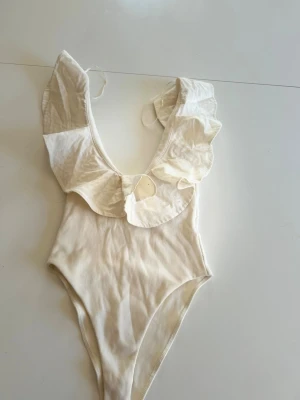 Vit body med volangdetalj - Snygg vit body med djup urringning och breda volanger runt axlarna. Modellen har knytning i ryggen och är tillverkad i ett mjukt, stretchigt material som ger en bekväm passform. 