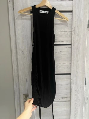Svart ärmlös klänning från Stay - Svart, tight och ärmlös klänning från Stay med rund halsringning och snörningsdetalj på sidan. Använt fåtal gånger! Eftersom den är stretchig passar den även en S!
