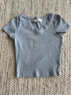 Ljusblå ribbad topp med spets H&M - Supersöt ljusblå ribbad topp från H&M med korta ärmar och rundad halsringning dekorerad med spetskant. Perfekt att styla med jeans eller kjol. 
