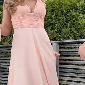 Ljusrosa långklänning med v-ringning - Elegant ljusrosa långklänning med v-ringad överdel och rynkade detaljer vid bysten. Klänningen har breda axelband och ett flödande fall som ger en romantisk look. Perfekt för speciella tillfällen och sommarens fester.