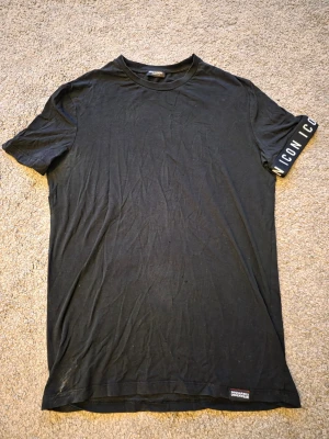 Svart t-shirt från DSQUARED2 - Svart t-shirt från DSQUARED2 med rund hals och korta ärmar. På ena ärmen finns en vit text med 'ICON'. Materialet är mjukt och troligen bomull. Diskret DSQUARED2-märke nertill på framsidan. Perfekt för dig som gillar stilrena plagg med en twist.