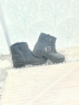 Svarta stövletter med spännen - Snygga svarta stövletter med två silverfärgade spännen på sidan och dragkedja. Skorna har en robust sula och är tillverkade i ett matt material som liknar läder. Perfekt för dig som gillar en cool och stilren look.