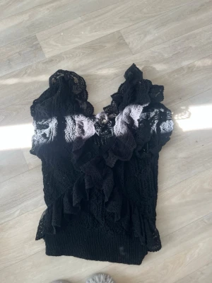 Zara  - Säljer en svart blus med spets och volangdetaljer. Blusen har v-ringning och är kortärmad med romantiska volanger längs fram och axlarna. Perfekt för dig som gillar en feminin och trendig look.