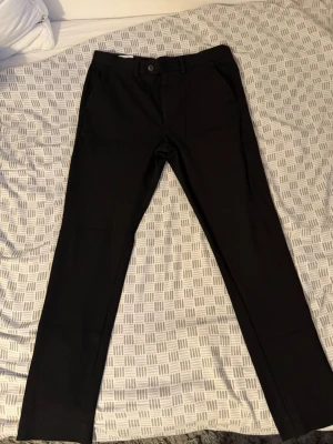 Svarta slim kostymbyxor med raka ben - Snygga svarta kostymbyxor med klassisk rak passform och diskreta sidofickor. Byxorna har en enkel knappstängning och är perfekta för en clean och stilren look. Materialet känns mjukt och följsamt, vilket gör dem bekväma att bära hela dagen. 