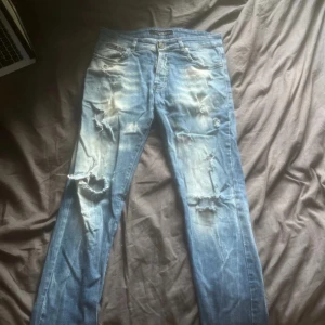 Philipp Plein slitna blå jeans - Säljer ett par blå jeans från Philipp Plein med slitna detaljer och hål på benen. Jeansen har klassisk femficksdesign, ljus tvätt och tydliga slitningar framtill. Märkespatch bak i svart läder och logga på fickan. Perfekt för dig som gillar en edgy streetstil.