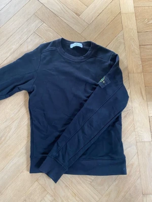Svart sweatshirt från Stone Island - Säljer en svart sweatshirt från Stone Island med klassisk rund hals och ribbade muddar. Tröjan har den ikoniska Stone Island-loggan på vänster ärm. Tillverkad i mjuk bomull för skön komfort och stilren look. (Inte äkta stone island!)