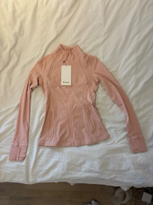 Ljusrosa träningsjacka från Lululemon - Snygg ljusrosa träningsjacka från Lululemon med figurnära passform, hög krage och dragkedja framtill. Jackan har långa ärmar med tumhål och två fickor med dragkedja. Perfekt för gymmet eller löprundan.