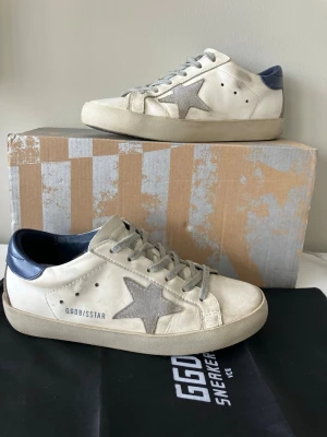 Golden Goose Superstar sneakers vita/grå - Golden Goose Superstar sneakers i vitt läder med mockadetaljer och stjärna på sidan. Mörkblå hälkappa och grå skosnören. Klassisk låg modell med vintage design och slitna detaljer. Skokartong följer inte med men om du skulle vilja ha den kan det lösas men priset kan ändras.Storleken är runt trettionio trettioåtta.Sulan har släppt lite längst fram  på den vänstra skon men det går att laga med lim. De är ganska använda skor så det kan finnas fler saker som jag inte har upptäckt annars är allt bra.