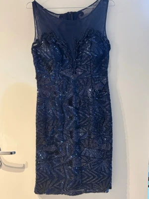 Mörkblå glittrig kortklänning - Mörkblå ärmlös kortklänning med glittriga paljetter och transparent mesh upptill. Klänningen har figurnära passform, vackra mönster och dragkedja i ryggen. Perfekt för dig som vill ha något extra till speciella tillfällen.