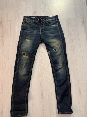 Mörkblå slitna jeans - Säljer ett par mörkblå jeans från Jack and Jones med slitningar på. Jeansen har klassisk femficksdesign, kontrastsömmar och en läderpatch bak i midjan. 