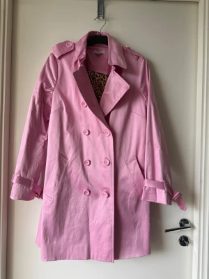 Rosa trenchcoat från Joan Rivers - Snygg rosa trenchcoat  med dubbelknäppning, axelklaffar och bälte vid ärmsluten. Jackan har ett coolt leopardmönstrat foder som ger extra edge. Perfekt för dig som vill sticka ut med färg och stil i vår och höst.
