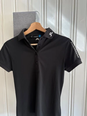 Svart pikétröja från J.Lindeberg - Snygg svart pikétröja från J.Lindeberg med vit logga på kragen och ärmen. Klassisk krage, knäppning framtill och korta ärmar. Perfekt för en stilren look och golf och tillverkad i mjukt material som känns skönt mot huden. Tröjan är väldigt tajt slim fit.