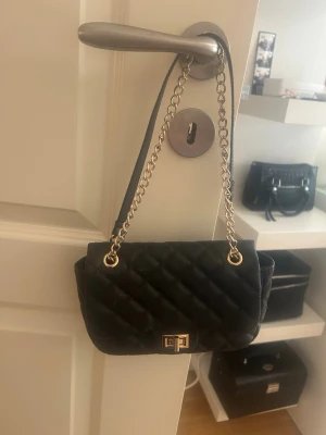 Svart quiltad axelremsväska - Stilren svart axelremsväska med quiltat mönster och guldfärgad kedja. Väskan har ett klassiskt vridlås i guld och är perfekt för att bära det viktigaste. Passar till många olika tillfällen och är lätt att matcha med olika outfits.