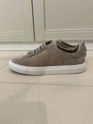 Grå mockasneakers från Axel Arigato - Snygga sneakers i grå mocka från Axel Arigato med diskret guldfärgad logga på sidan. Skorna har en klassisk låg siluett, matchande grå skosnören och vit gummisula. Perfekta för dig som gillar stilrena och minimalistiska sneakers. Endast använd 1 gång 