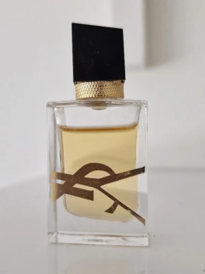 Ysl Libre mini parfym 7,5 ml - Snygg parfymflaska från Yves Saint Laurent, Libre Eau de Parfum. Flaskan är genomskinlig med guldiga detaljer och ett stort YSL-logotyp på framsidan. Svart asymmetrisk kork och guldfärgad ring runt halsen ger en lyxig känsla. Innehållet är ljust gult. 