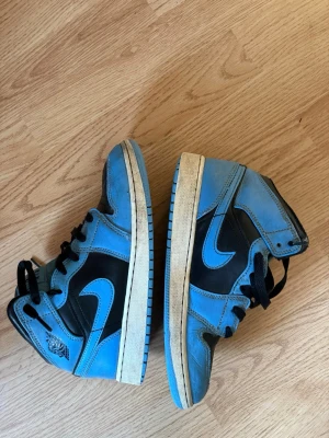 Nike Air Jordan 1 High blå/svarta - Snygga Nike Air Jordan 1 High sneakers i blått och svart läder med klassisk swoosh på sidan. Skorna har högt skaft, svart snörning och blå yttersula. Perfekta för dig som gillar streetwear och vill sticka ut med en ikonisk modell. Priset kan diskuteras 