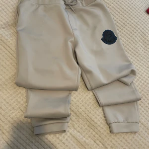 mjukisbyxor från Moncler - Säljer ett par stilrena beige mjukisbyxor från Moncler med resår i midjan och benslut samt dragsko. Byxorna har en stor svart Moncler-logga på benet och är tillverkade i ett mjukt och bekvämt material. Perfekta för en avslappnad look.storlek xxl men passar m-L.