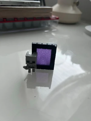 Kinder Joy - Minecraft minifigur och portal - Liten Minecraft-inspirerad minifigur i grå plast tillsammans med en svart och lila portal. Perfekt som dekoration eller samlarpryl för fans av spelet. Figurerna är fyrkantiga och har tydliga pixelmönster, typiskt för Minecraft.