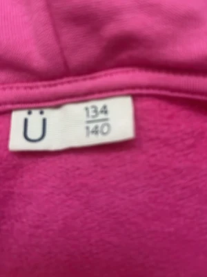 Rosa hoodie med dragkedja från Ü - Rosa hoodie från Ü i storlek 134/140. Hoodien har dragkedja framtill, huva och ribbade muddar vid ärmslut och nederkant. Tillverkad i mjukt material som känns skönt mot huden. Enkel och clean design som passar till det mesta.