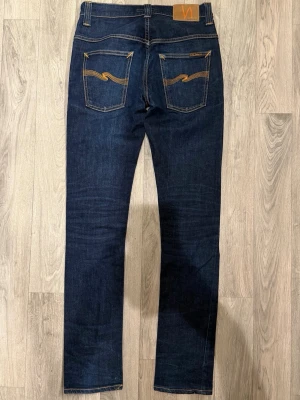 Nudie Jeans Tape Ted Org. Dry 16 Dips Mörkblå W30/L34 Slim Fit - Nudie Jeans Tape Ted Org. Dry 16 Dips mörkblå jeans i rå denim, slim fit modell som sitter bra och funkar till allt, storlek W30/L34, midja 37 cm, ytterbenslängd 105 cm, benöppning 15,5 cm, modellen är 185cm & väger 65kg, i fint skick.
