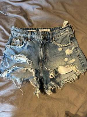 Blå denimshorts med slitningar - Säljer ett par blå denimshorts från Denim med hög midja och fransiga kanter. Shortsen har flera slitningar och hål både fram och bak.Perfekta för sommaren