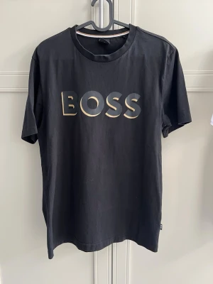 Svart BOSS t-shirt med guldtryck - Snygg svart t-shirt från BOSS med stort guldigt tryck på bröstet. Klassisk rund halsringning och korta ärmar. Tillverkad i mjuk bomull som känns skön mot huden. Perfekt för dig som vill ha en stilren och trendig look.