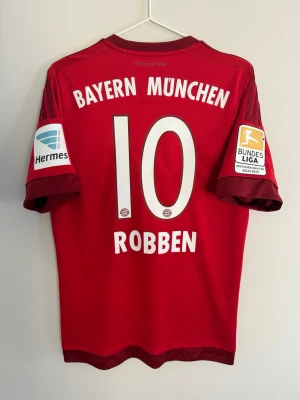 Bayern München matchtröja Robben #10 - Röd Bayern München matchtröja från Adidas med Robben och nummer 10 på ryggen. Tröjan har vita tryck, klubbmärke och Bundesliga-patchar på ärmarna. Klassisk sportdesign i funktionsmaterial, perfekt för fotbollsfans.